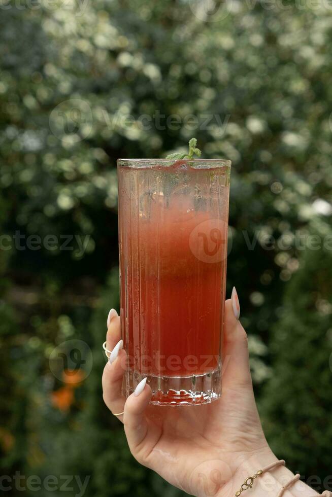 Lemonade with ice in female hand on greenery background photo