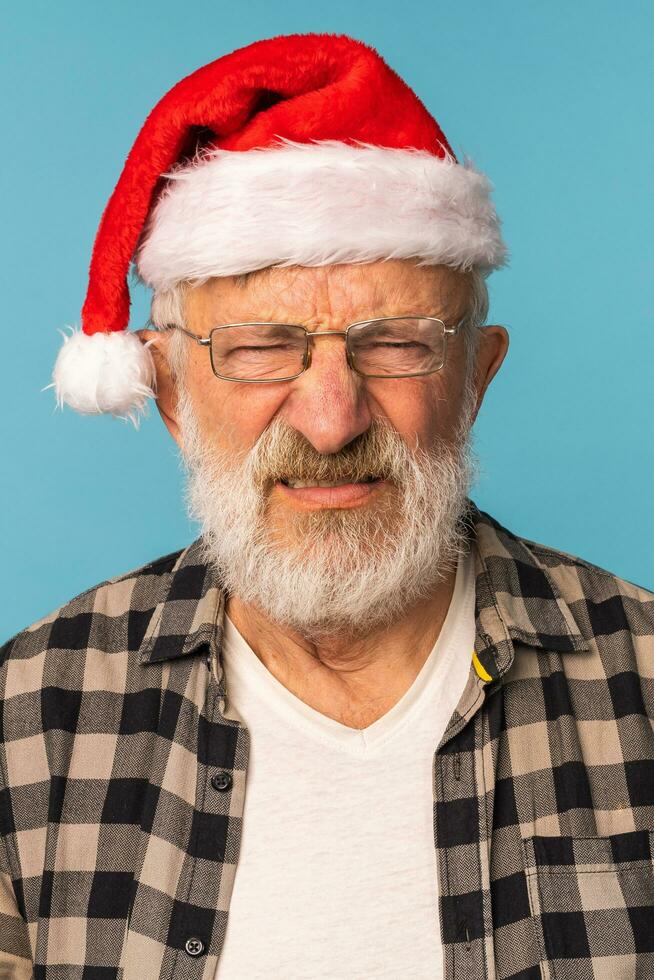 estudio retrato de blanco barba antiguo hombre en Papa Noel sombrero mirando a cámara con triste enojado expresión - emoción y malo Navidad estado animico concepto foto