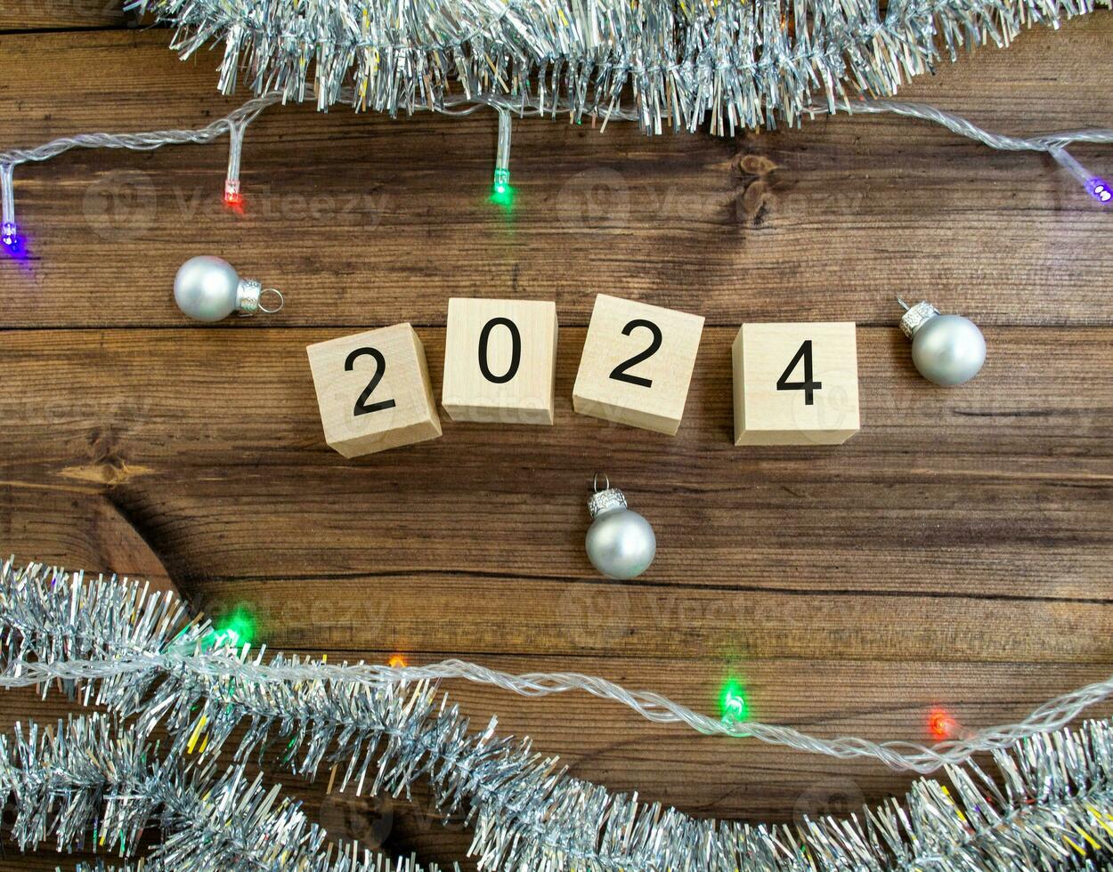 New Year's card 2024. Wooden cubes with numbers 2024, tinsel, garland and New Year's balls on a wooden background. photo