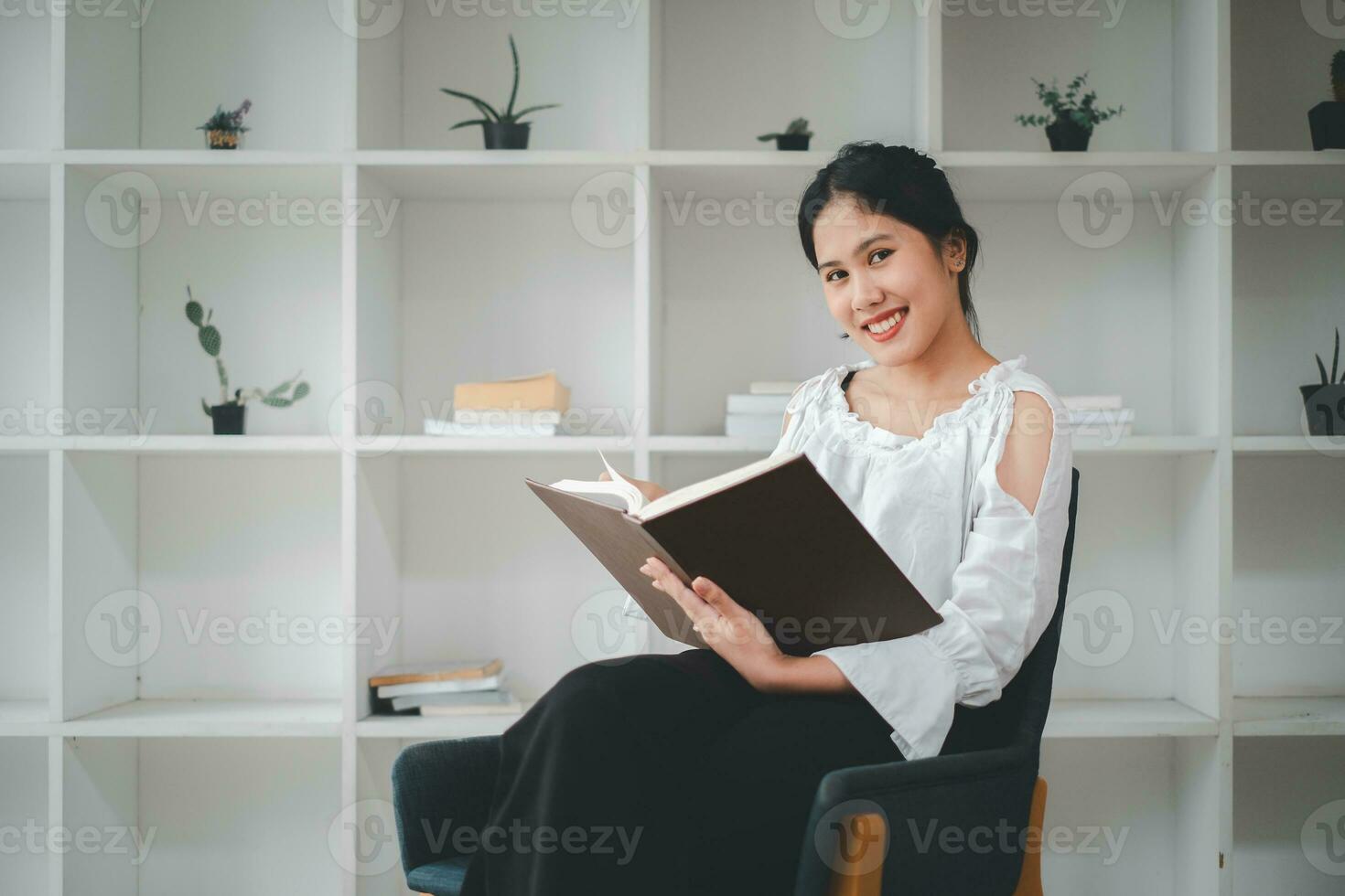 Side view of asian woman sitting armchair with open book and reading in living room. photo