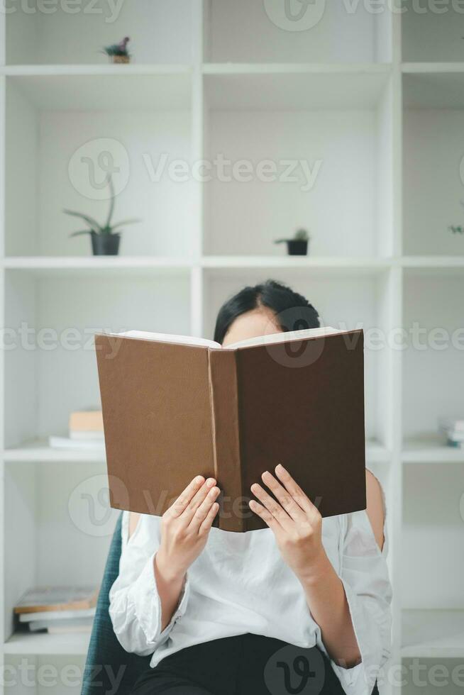 asiático mujer sentado en acogedor Sillón con abierto libro adentro. hermosa joven dama leyendo emocionante historia, disfrutando perezoso mañana, foto