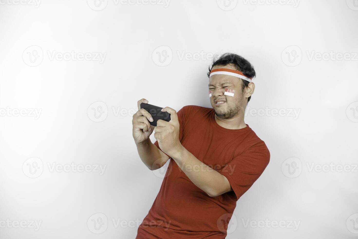 A portrait of a shocked Asian man wearing headband, holding his phone while his mouth wide open, isolated by white background. Indonesia's independence day concept photo