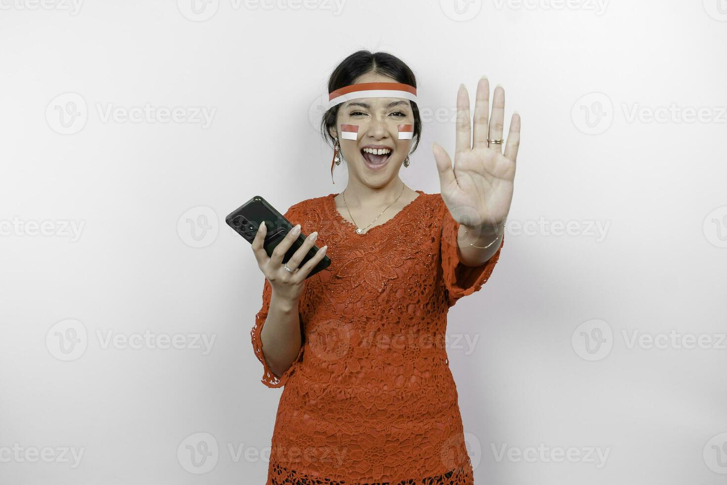 Attractive Asian woman wearing red kebaya and headband with hand gesture pose rejection or prohibition while holding her phone isolated by white background photo