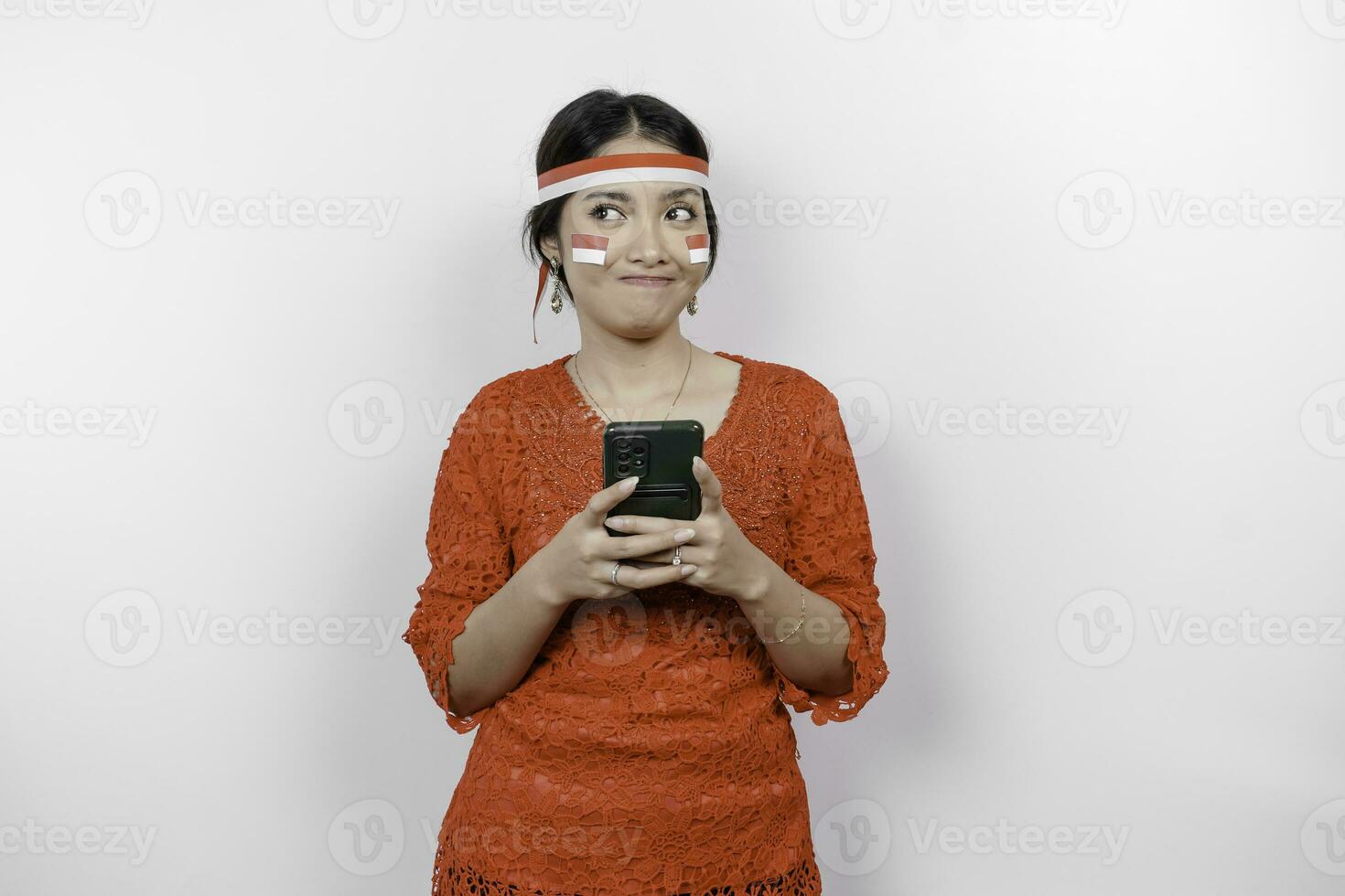 A smart young woman wearing headband and red kebaya is looking aside to an idea on copy space while holding her phone, isolated by white background. Indonesia's independence day. photo