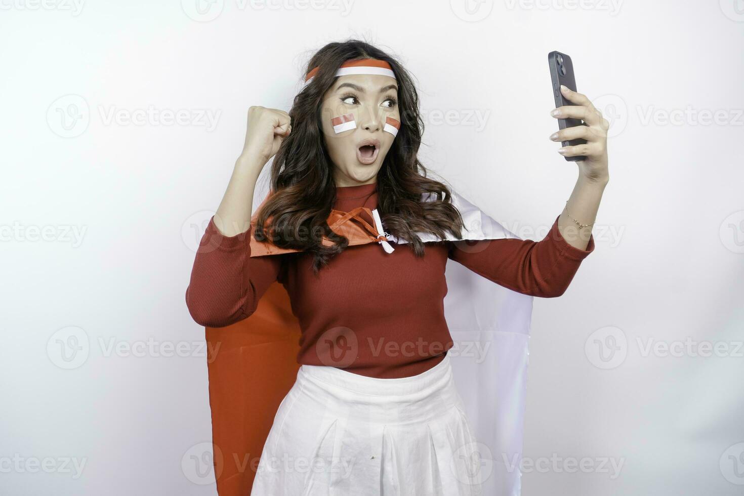 A young Asian woman with a happy successful expression while holding her phone and wearing red top, flag headband and cape isolated by white background. Indonesia's independence day concept. photo