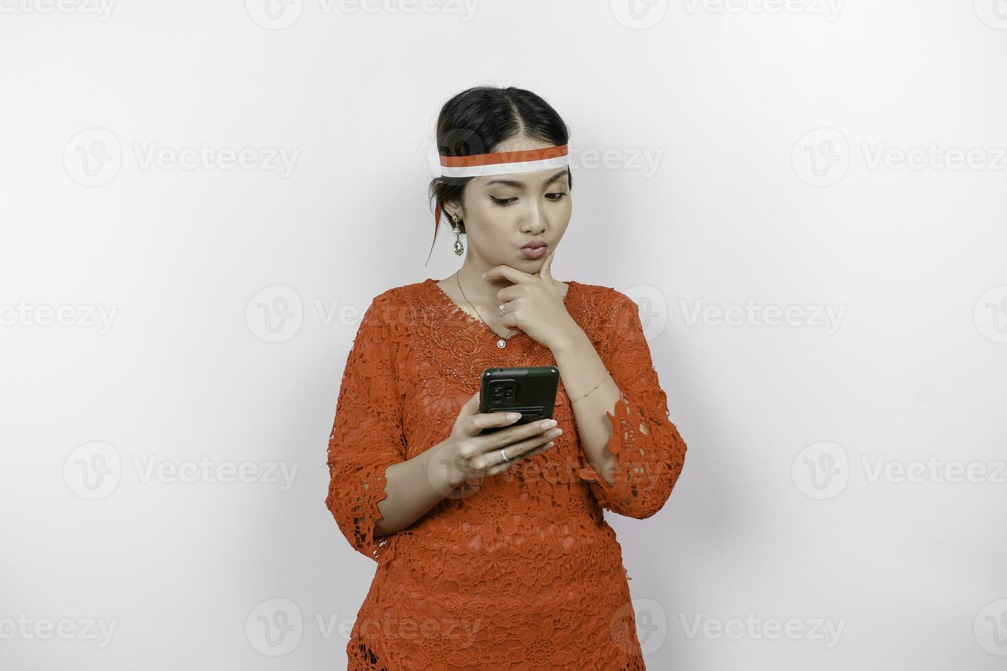 A smart young woman wearing headband and red kebaya is looking aside to an idea on copy space while holding her phone, isolated by white background. Indonesia's independence day. photo