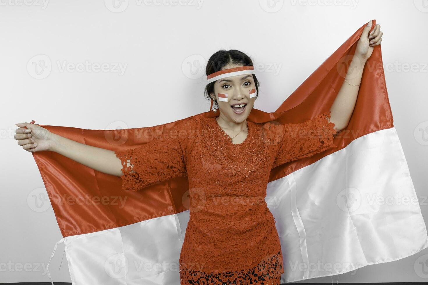 Happy smiling Indonesian woman wearing red kebaya and headband holding Indonesia's flag to celebrate Indonesia Independence Day isolated over white background. photo