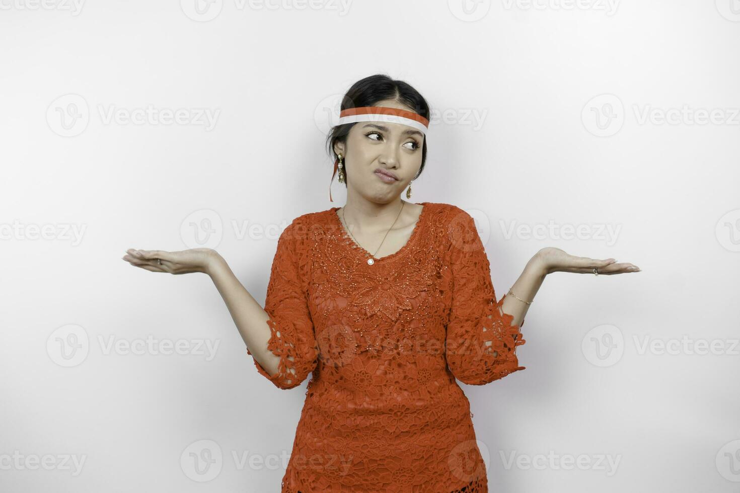 A thoughtful young woman wearing headband and red kebaya is looking aside confuse between choices, isolated by white background. Indonesia's independence day. photo