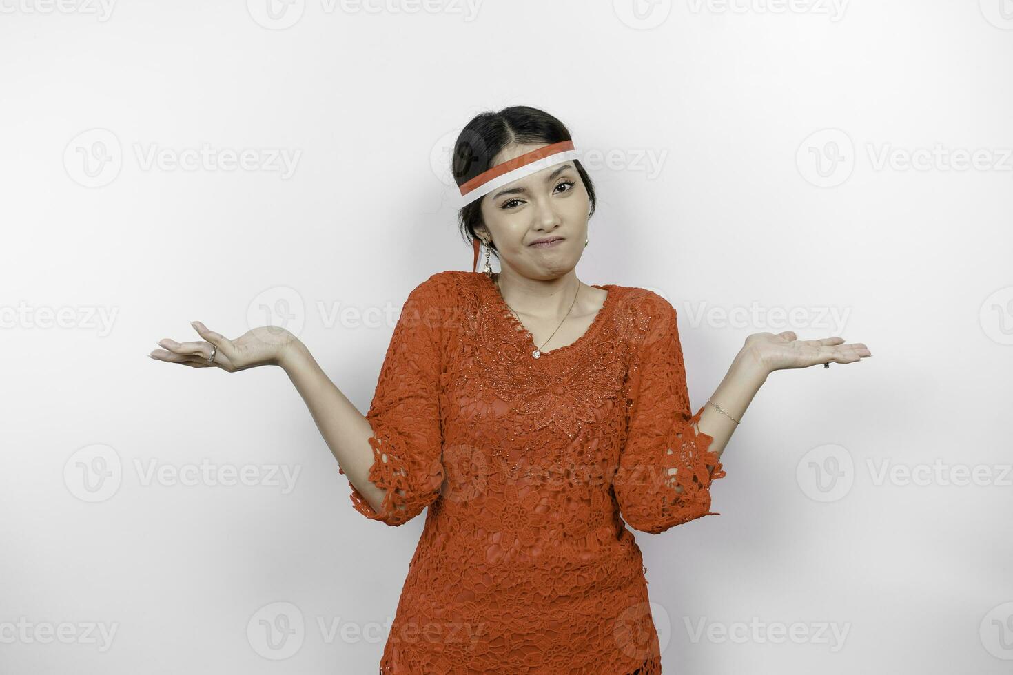 A thoughtful young woman wearing headband and red kebaya is looking aside confuse between choices, isolated by white background. Indonesia's independence day. photo