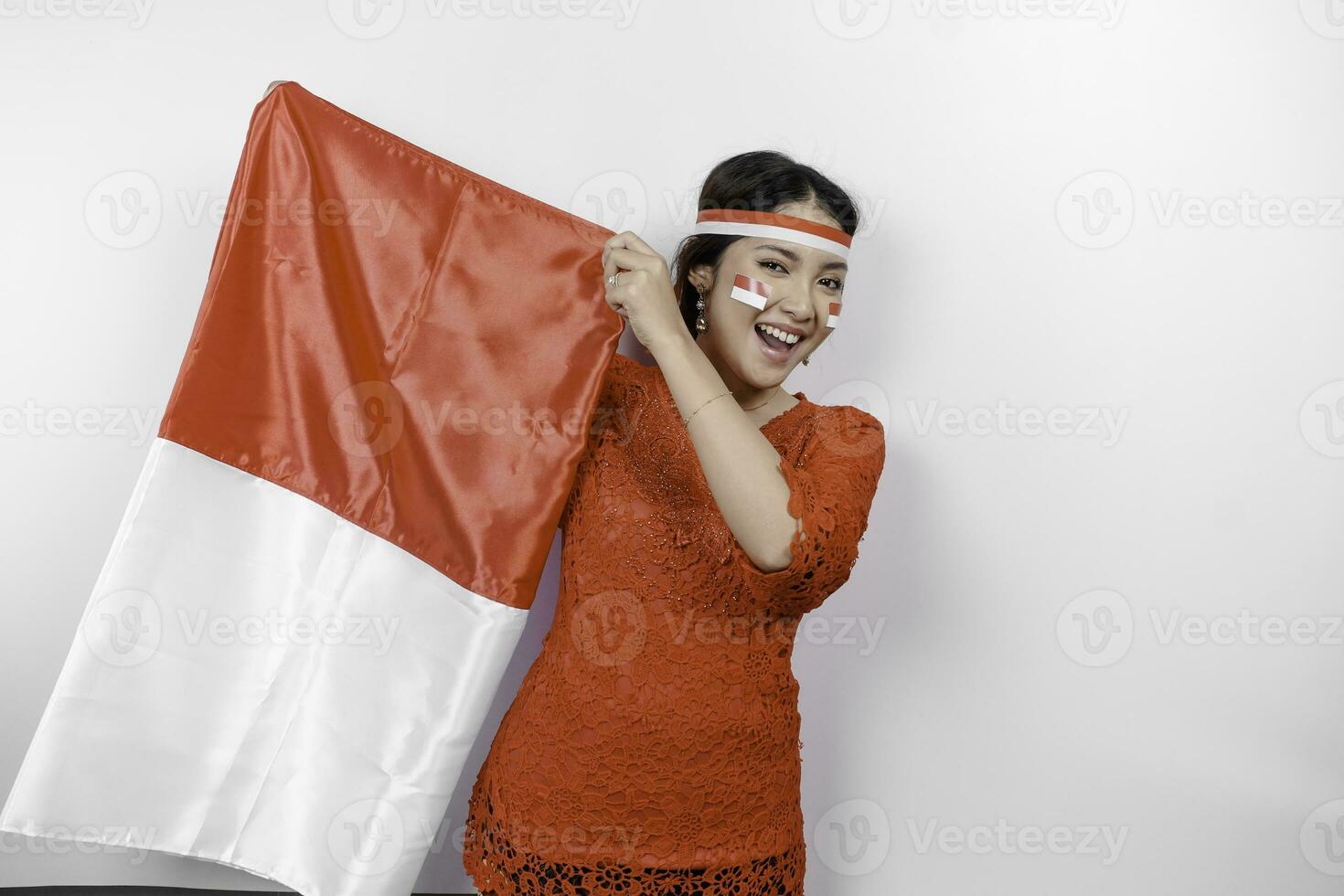 Happy smiling Indonesian woman wearing red kebaya and headband holding Indonesia's flag to celebrate Indonesia Independence Day isolated over white background. photo