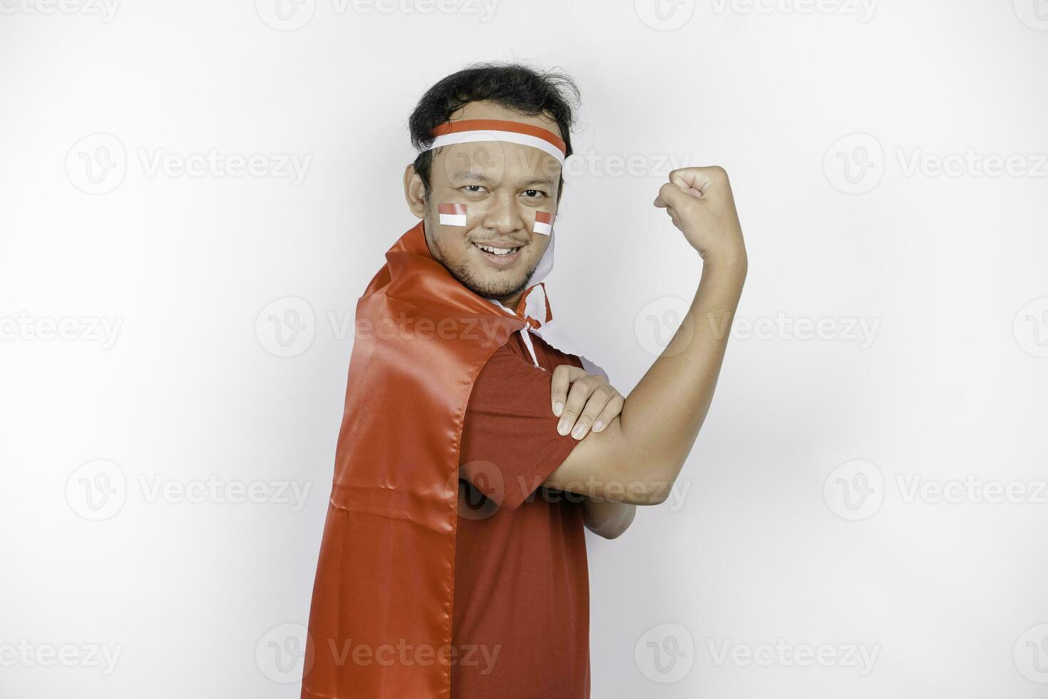 Excited Asian man wearing a red top, flag cape and headband, showing strong gesture by lifting his arms and muscles smiling proudly. Indonesia's independence day concept. photo