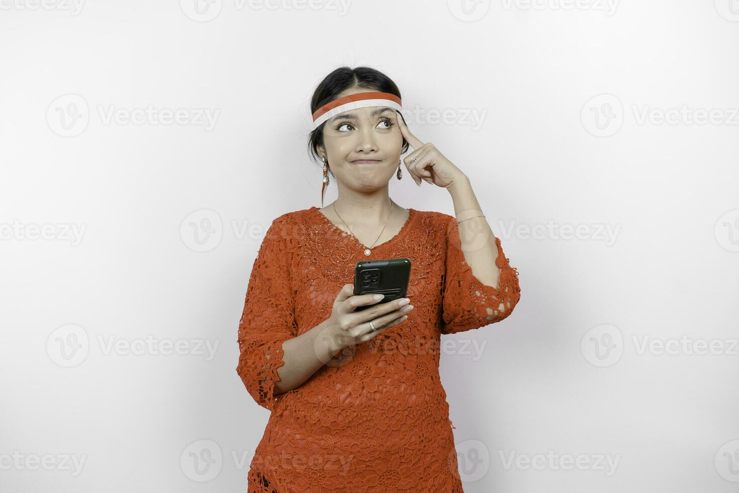 A smart young woman wearing headband and red kebaya is looking aside to an idea on copy space while holding her phone, isolated by white background. Indonesia's independence day. photo