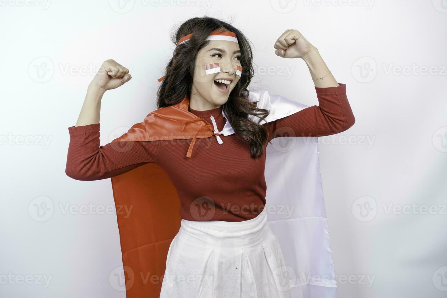Excited Asian woman wearing a red top, flag cape and headband, showing strong gesture by lifting her arms and muscles smiling proudly. Indonesia's independence day concept. photo