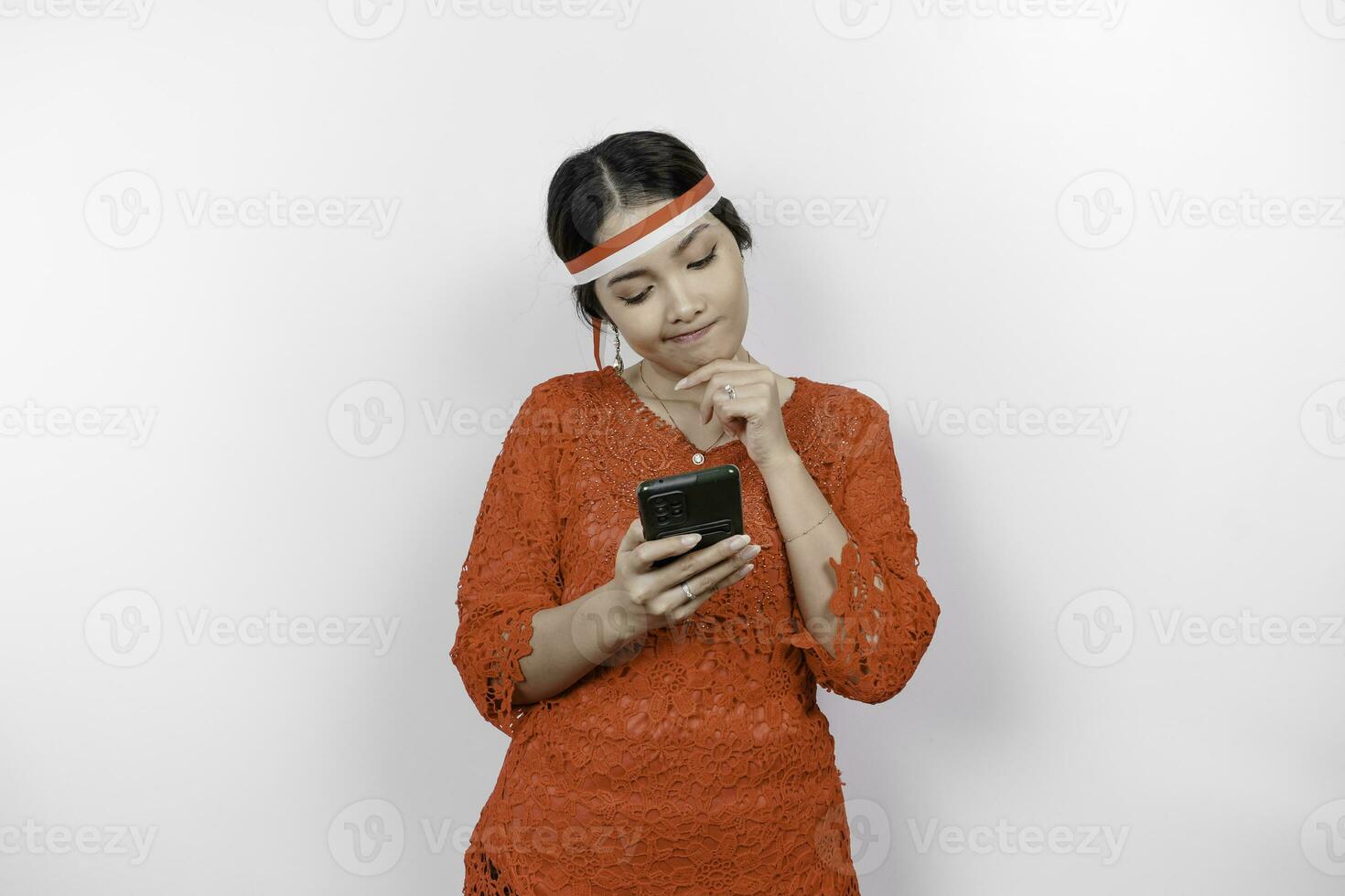 A smart young woman wearing headband and red kebaya is looking aside to an idea on copy space while holding her phone, isolated by white background. Indonesia's independence day. photo