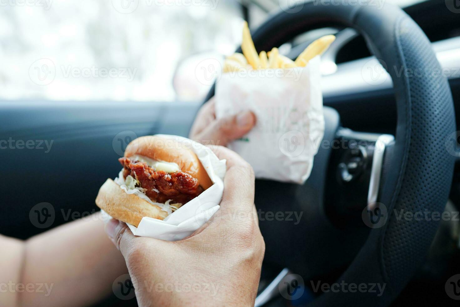 Asian woman driver hold and eat hamburger and french fries in car, dangerous and risk an accident. photo