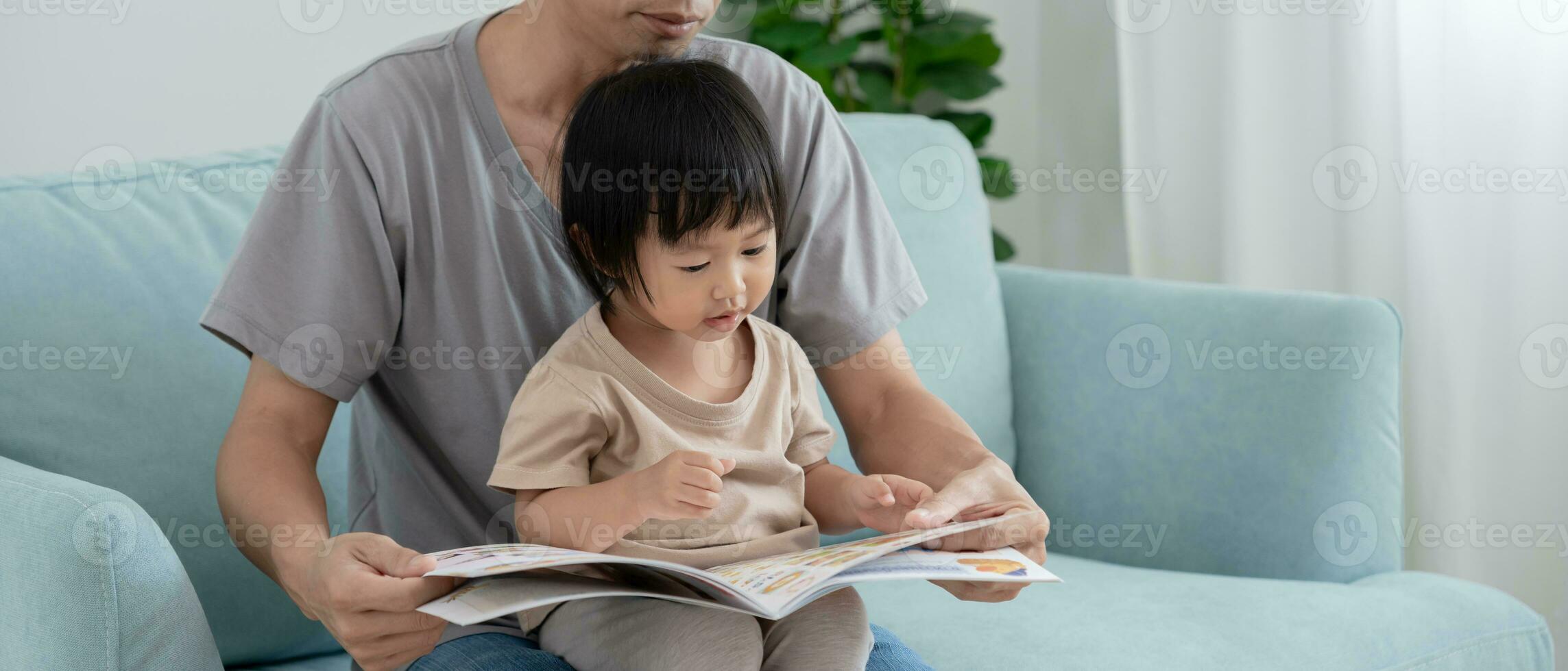 Asian father relax and read book with baby time together at home. parent sit on sofa with daughter and reading a story. learn development, childcare, laughing, education, storytelling, practice. photo