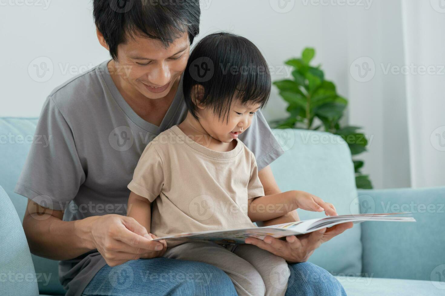 Happy Asian father relax and read book with baby time together at home. parent sit on sofa with daughter and reading a story. learn development, childcare, laughing, education, storytelling, practice. photo
