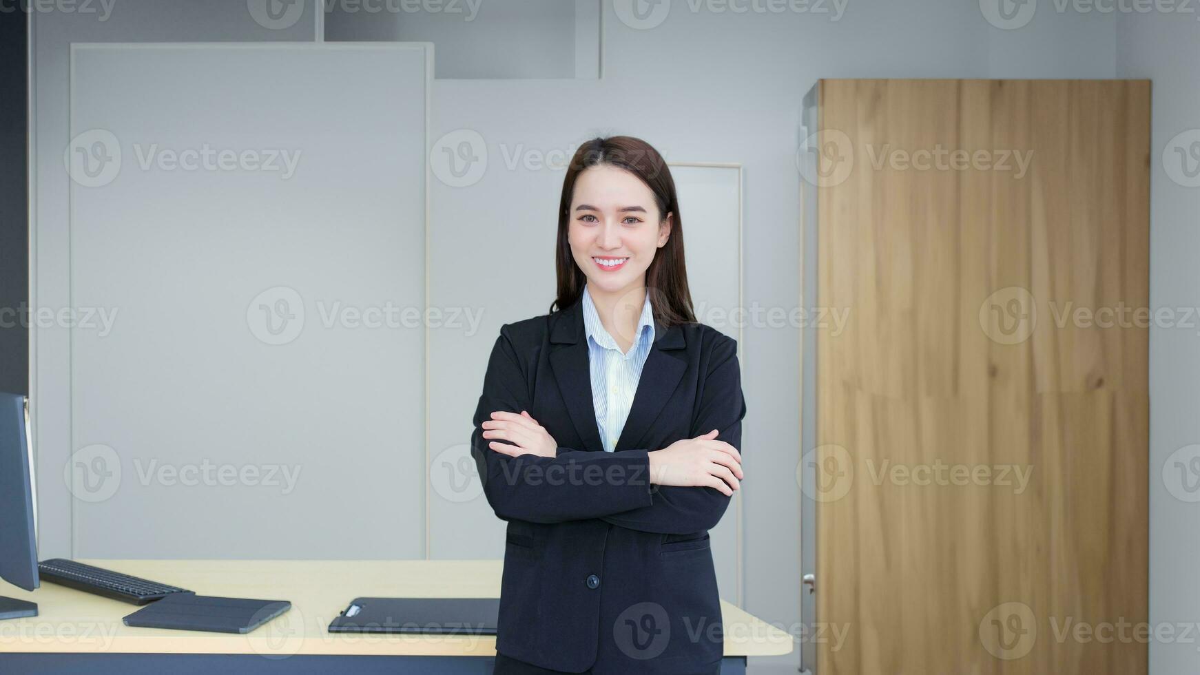mujer trabajadora profesional asiática que tiene el pelo largo viste traje formal negro con camisa azul mientras cruza el brazo y sonríe confiada en la oficina. foto