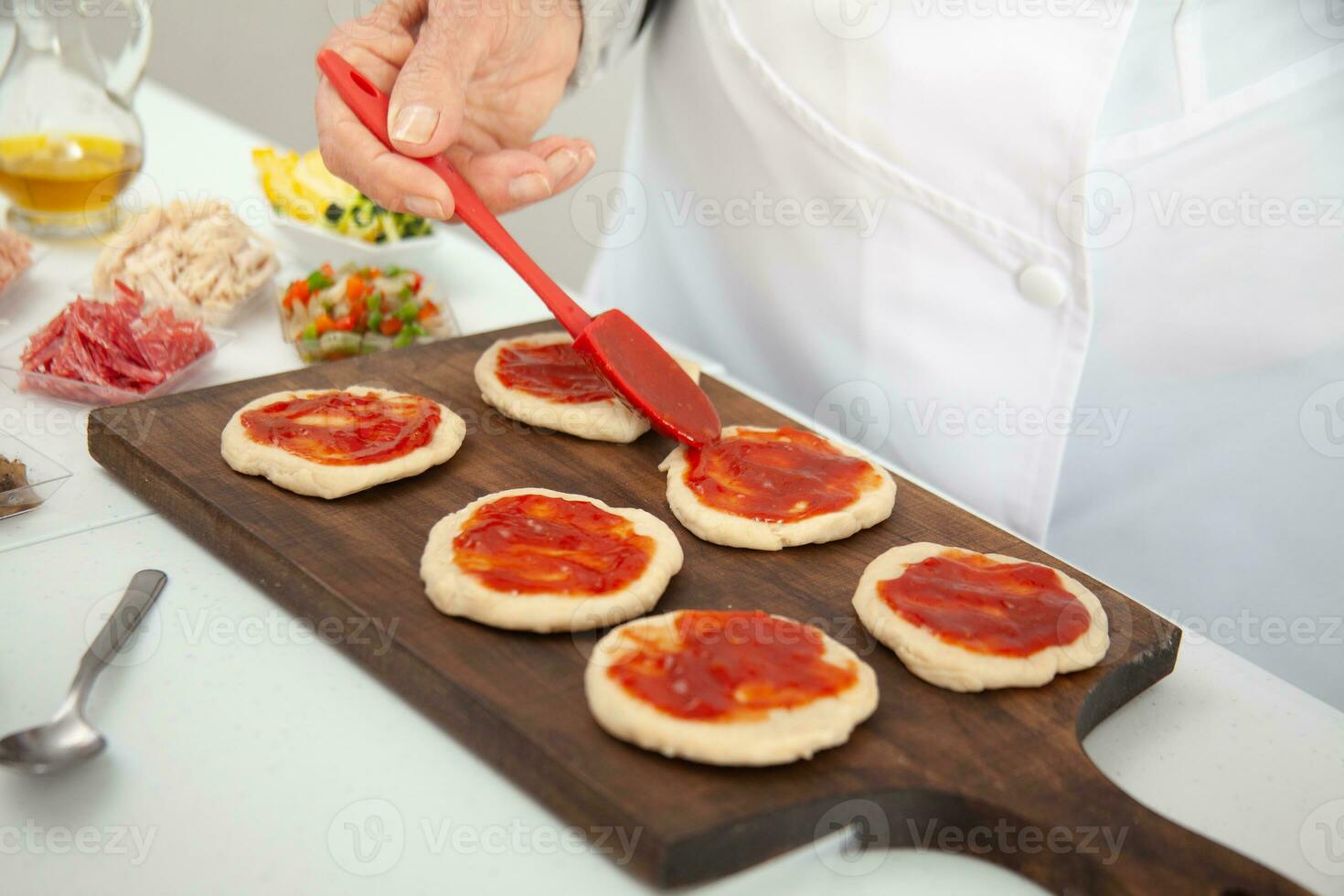 Senior woman adding pizza sauce to the stretched dough. Delicious homemade mini pizzas preparation. photo