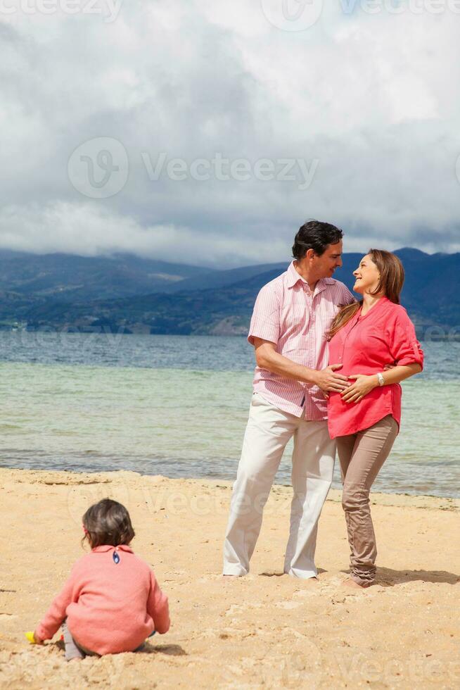 Young couple waiting for their second baby having fun with their baby girl at the beautiful white beach of Lake Tota located in the department of Boyaca at 3,015 meters above sea level in Colombia photo