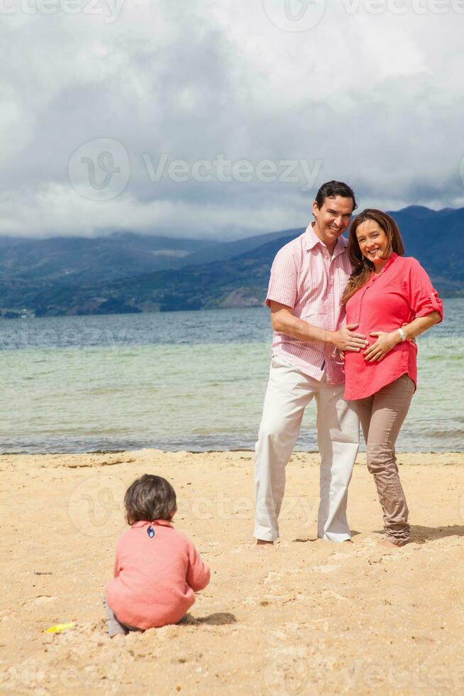 Young couple waiting for their second baby having fun with their baby girl at the beautiful white beach of Lake Tota located in the department of Boyaca at 3,015 meters above sea level in Colombia photo