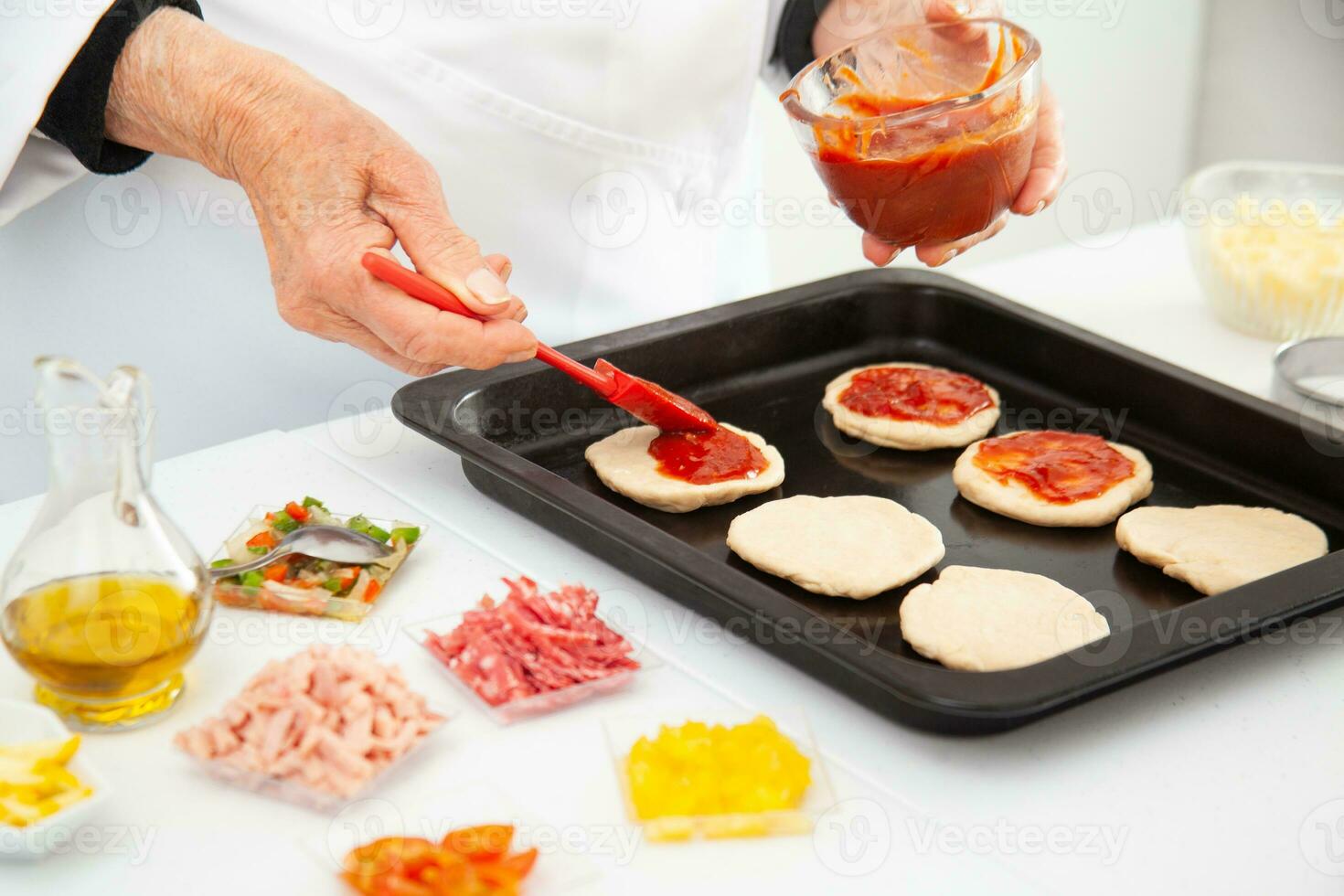 Senior woman adding pizza sauce to the stretched dough. Delicious homemade mini pizzas preparation. photo