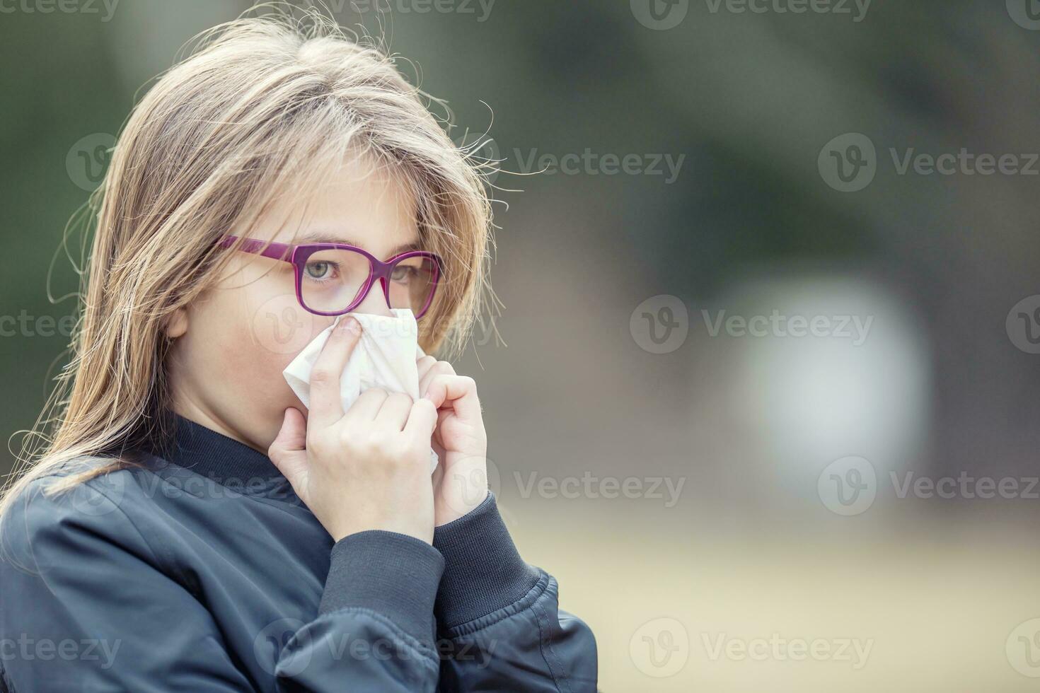 Girl with allergy symptom blowing nose. Teen girl using a tissue in a park. photo