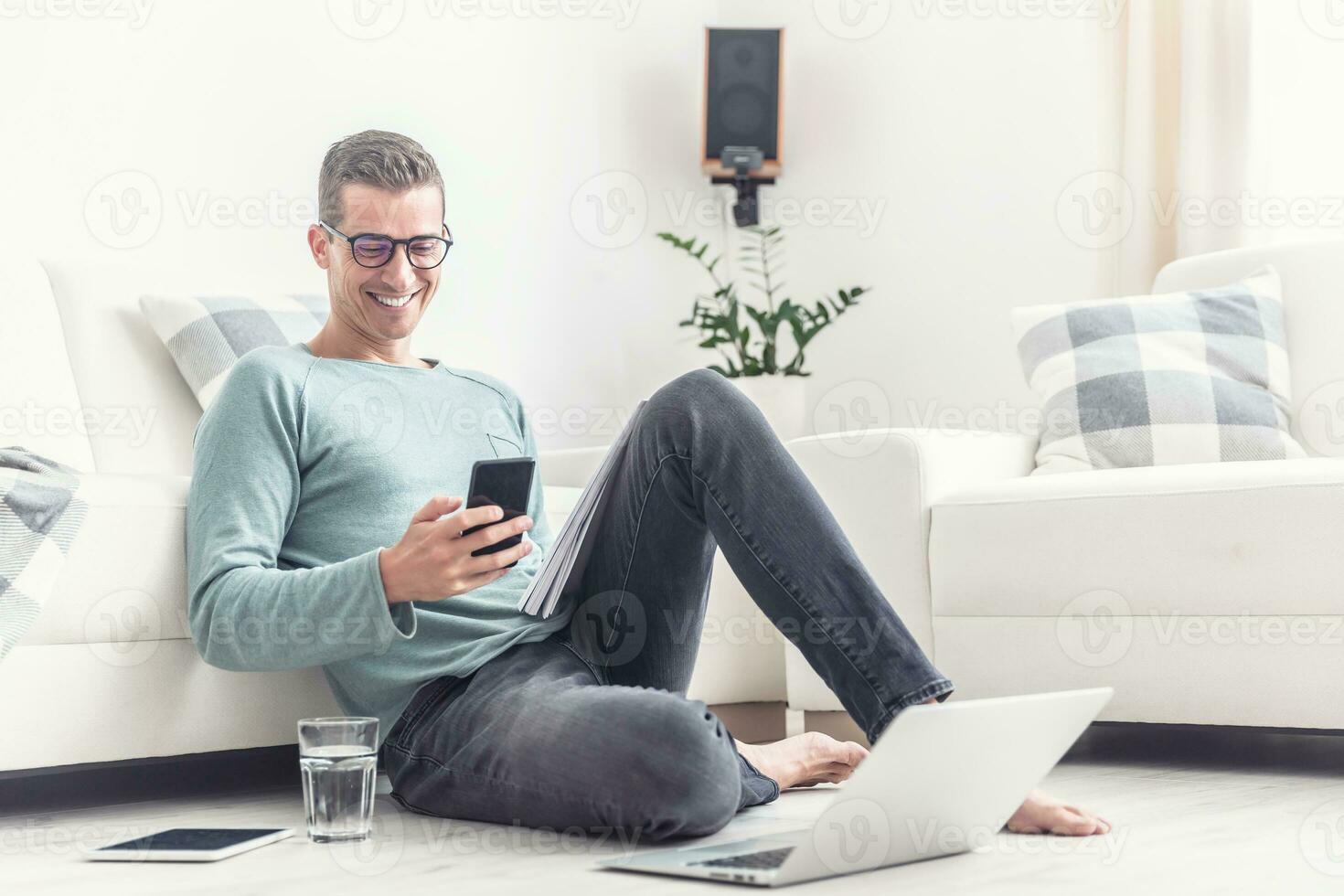 Smiling man texts on his mobile phone during his home office hours at home, sitting on a living room floor photo