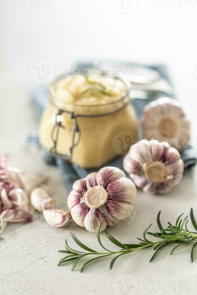 Homegrown garluc bulbs in front of a glass jar full of fresh garlic paste laid on kitchen cloth with rosemary photo