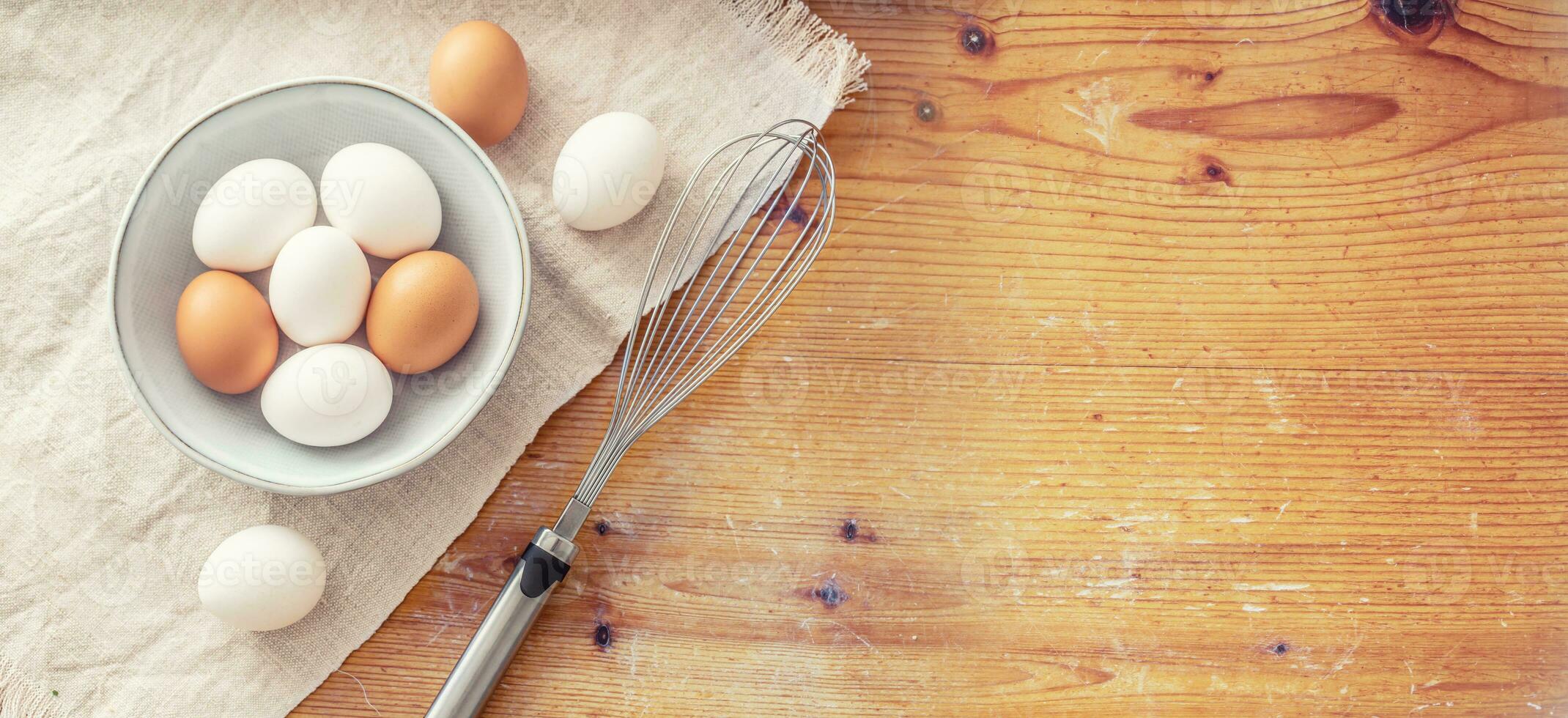 Rustic cutting board and tablecloth with nine eggs in and out of a bown and a metal steel whisk photo