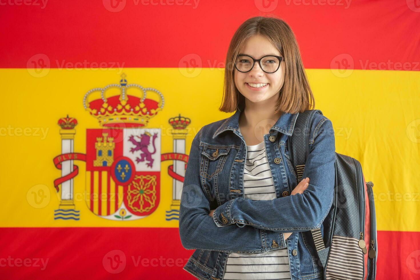 Teenage girl in glasses wears backpack and stands smiling in front of flag of Spain photo