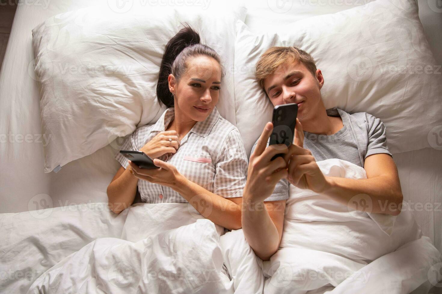 Top view of a couple sharing interesting and funny things on their mobile phones while lying in white bedding before sleep photo