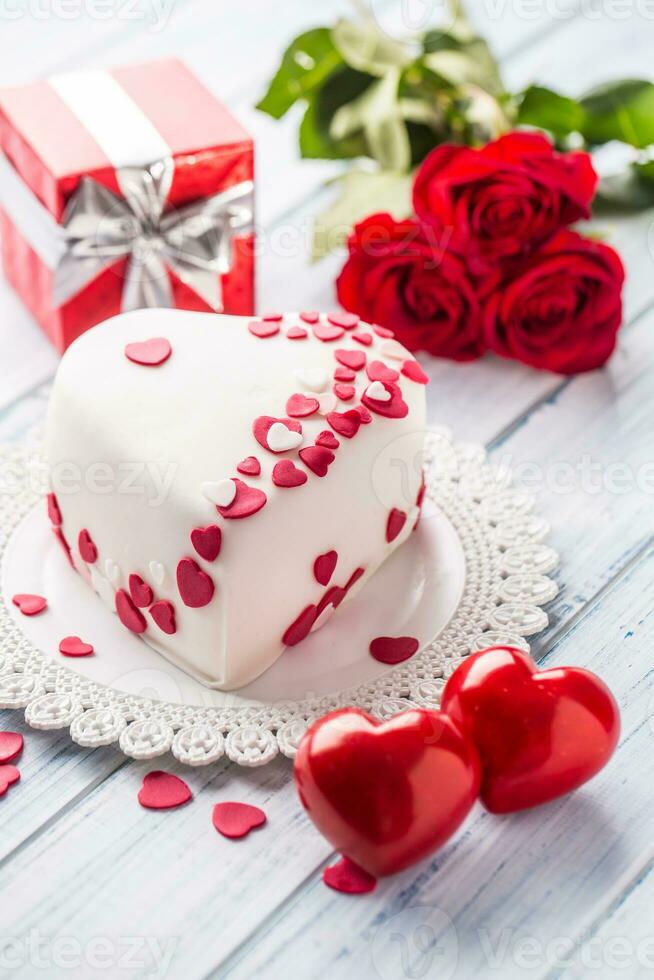 Marzipan white cake in the shape of a heart with red hearts. As the decoration bouquet of red roses a gift from the ribbon. Wedding or valentines day concept. photo