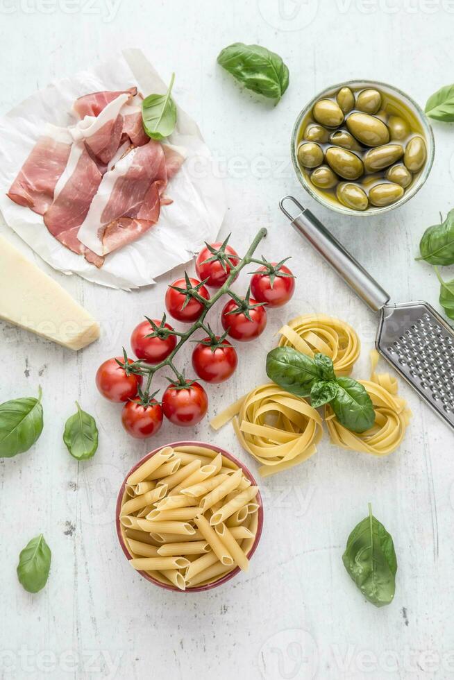 Italian or mediterranean food cuisine and ingredients on white concrete table. Tagliatelle pene pasta  olives olive oil tomatoes parmesan cheese prosciutto and basil leaves on white concrete board photo