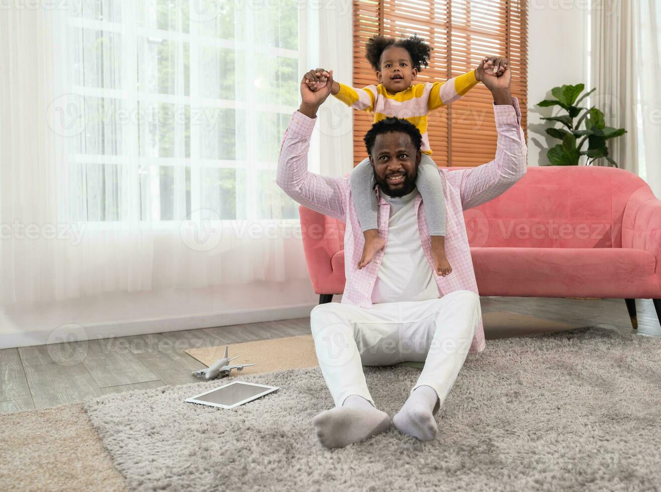 Curly little daughter riding father neck with open hands and having fun together in living room. photo