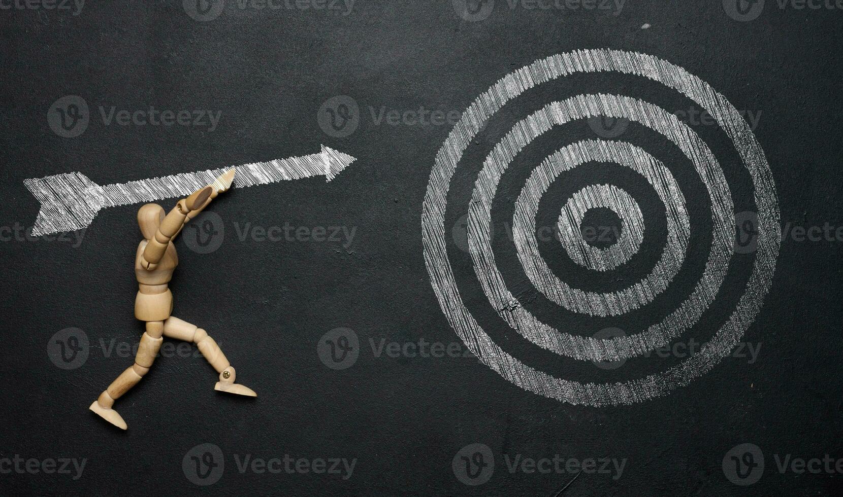 A wooden mannequin holds a drawn arrow and aims precisely at the target on a black chalkboard. The concept of achieving set goals photo