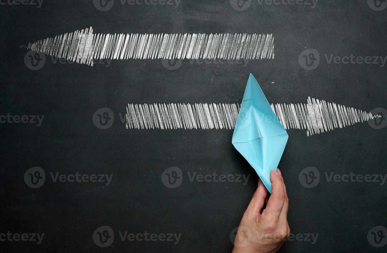 un mujer mano sostiene un azul papel barco en un negro tiza tablero y flechas en diferente direcciones, el elección de el dirección de movimiento foto