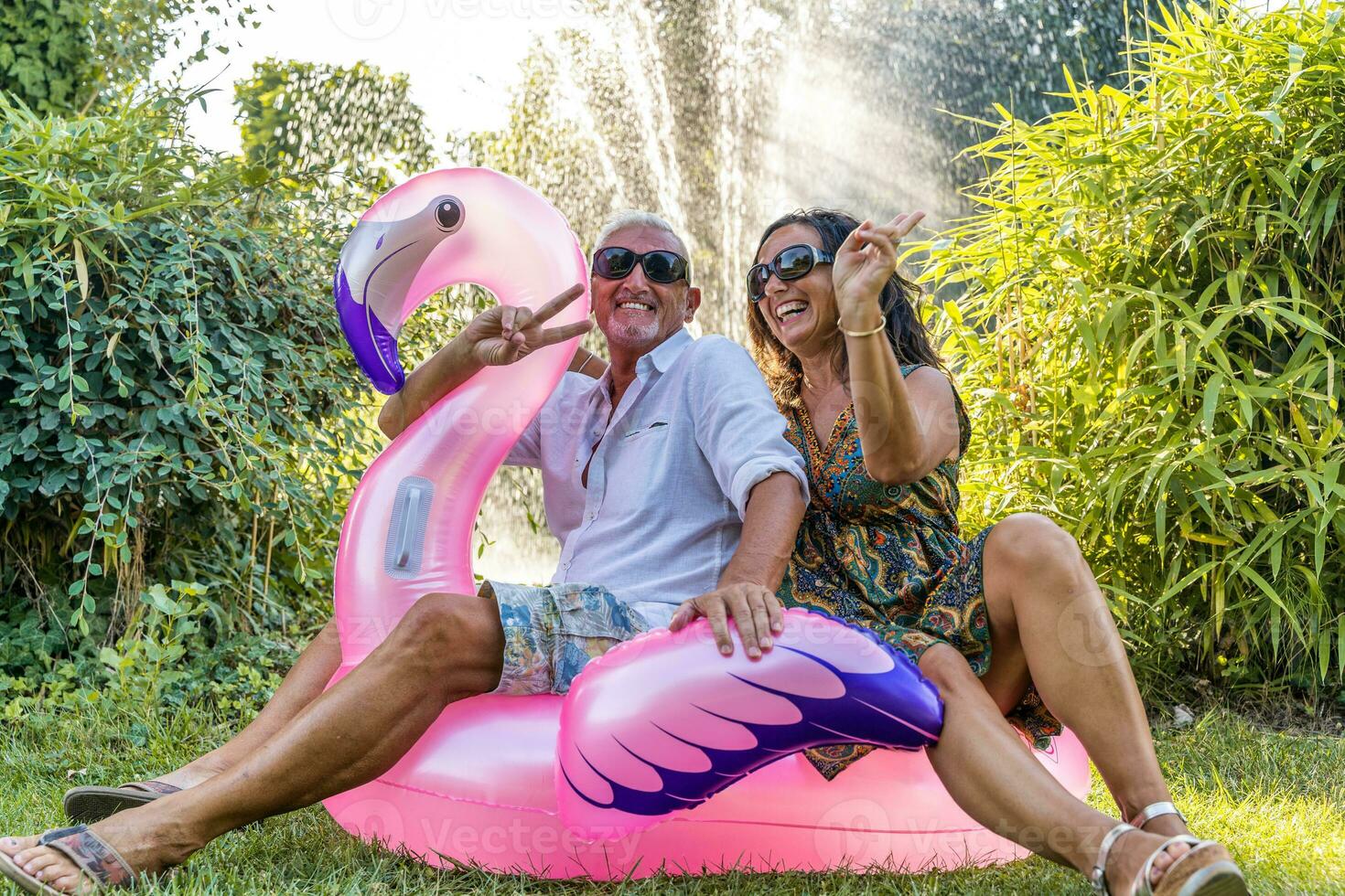 beautiful smiling middle aged couple sitting on pink flamingo inflatable toy photo
