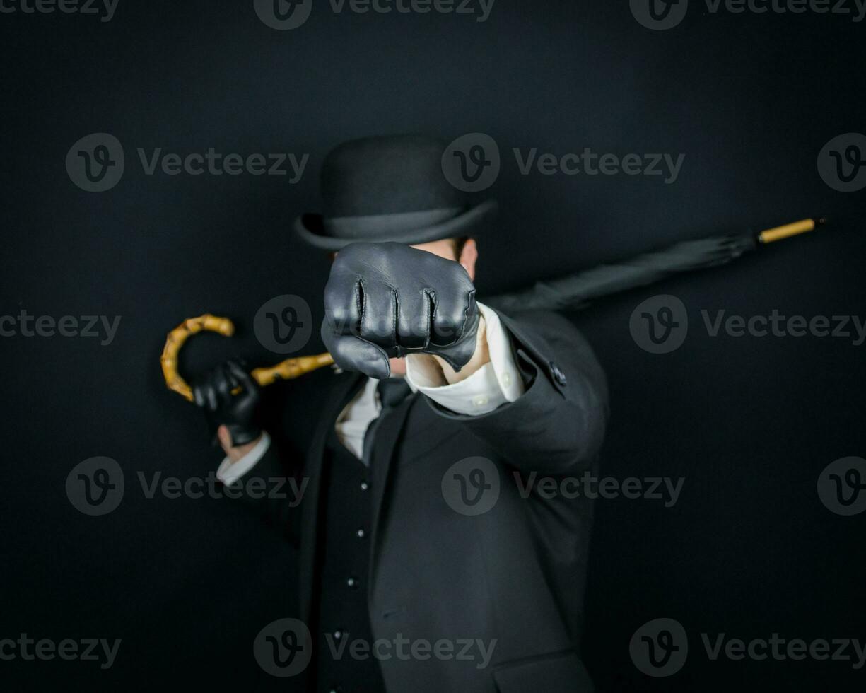 retrato de Caballero en oscuro traje y jugador de bolos sombrero participación un paraguas y lanzamiento un puñetazo. Clásico Inglés Caballero hombre de acción. foto