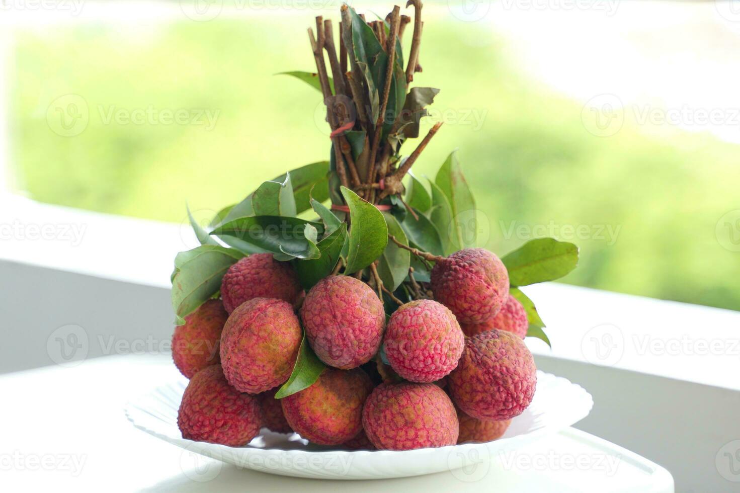 A plate of lychee bunches on a white table, a blurred background of green trees. They are reddish pink or bright red. seed coat Milky white that is full of water. photo