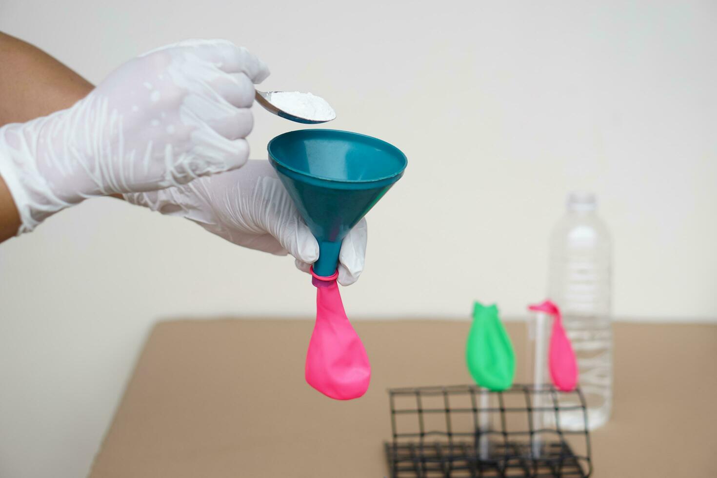 Closeup hands wears gloves holds funnel and flat pink balloon, put spoon of baking soda powder to pour into balloon. Concept, science experiment about reaction of chemical substance . photo