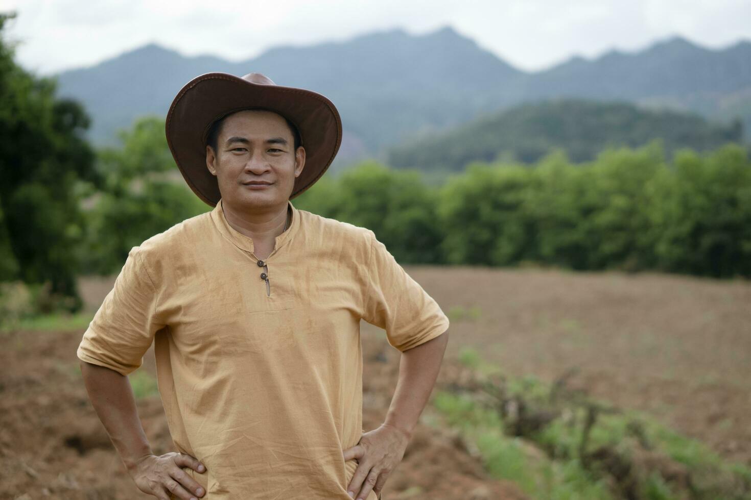 hermoso asiático hombre granjero usa sombrero, marrón camisa, poner manos en cintura, siente seguro, soportes a agricultura tierra antes de creciente cultivos. concepto, agricultura ocupación. tailandés granjero estilo de vida. foto