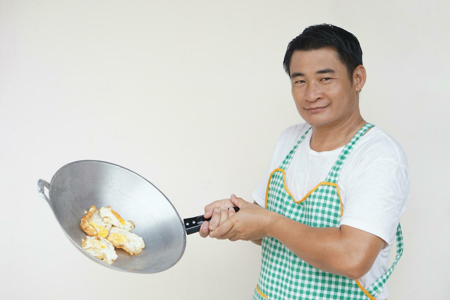 Handsome Asian man is cooking fried eggs, wears white shirt and apron, holds frying pan and ladle spatula . Concept, love cooking. Kitchen lifestyle. photo