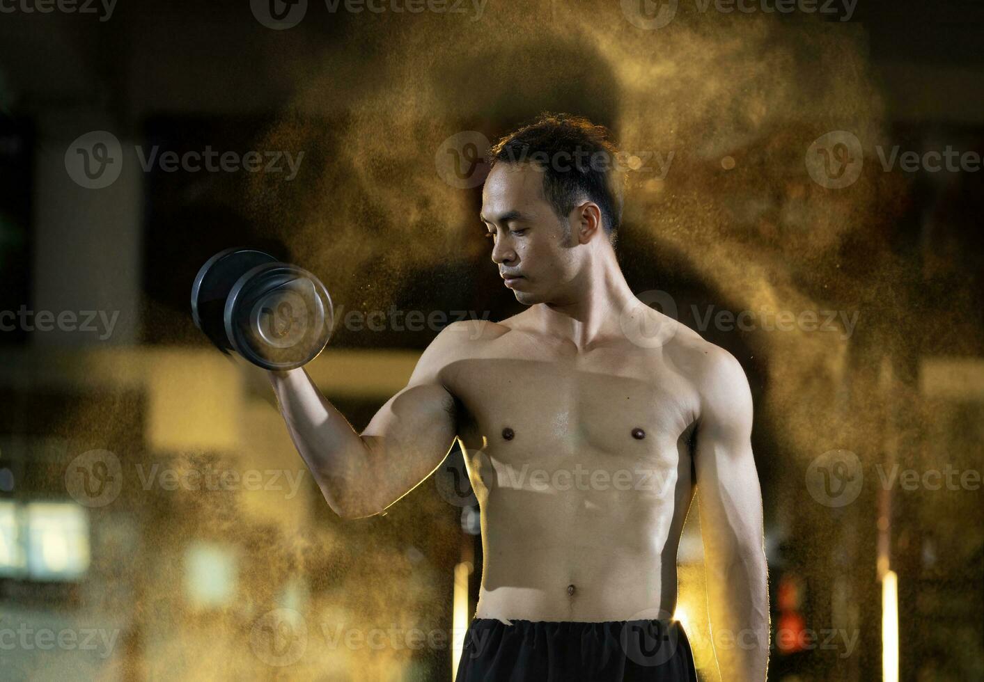 Asian muscular shirtless sport man is practice weight training on dumbbell for biceps and triceps muscle inside gym with dark backgroun and light smoke glow for exercising and workout concept photo