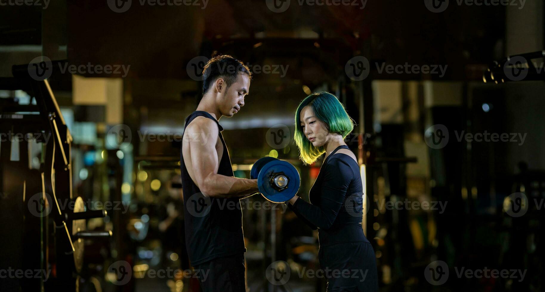 Asian woman is using easy bar as beginner in weight training on barbell for arm and core muscle inside gym with support from trainer to prevent injury dark background for exercising and workout photo