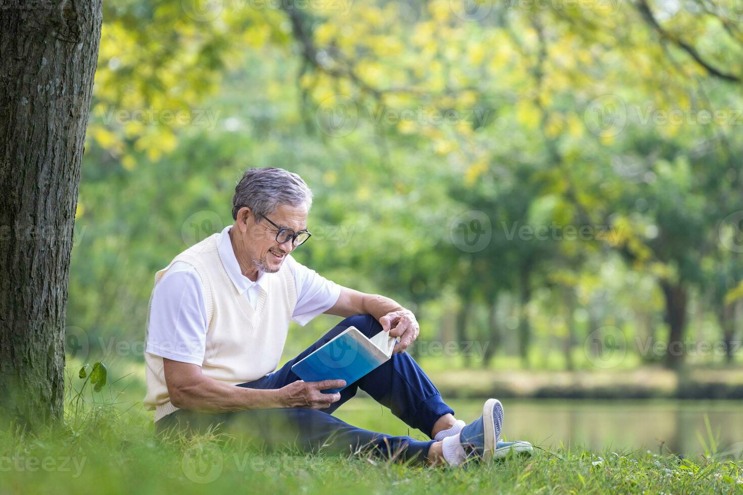 Senior Asian man reading a book while sitting under the tree by the lake at the public park for recreation, leisure and relaxation in nature concept photo
