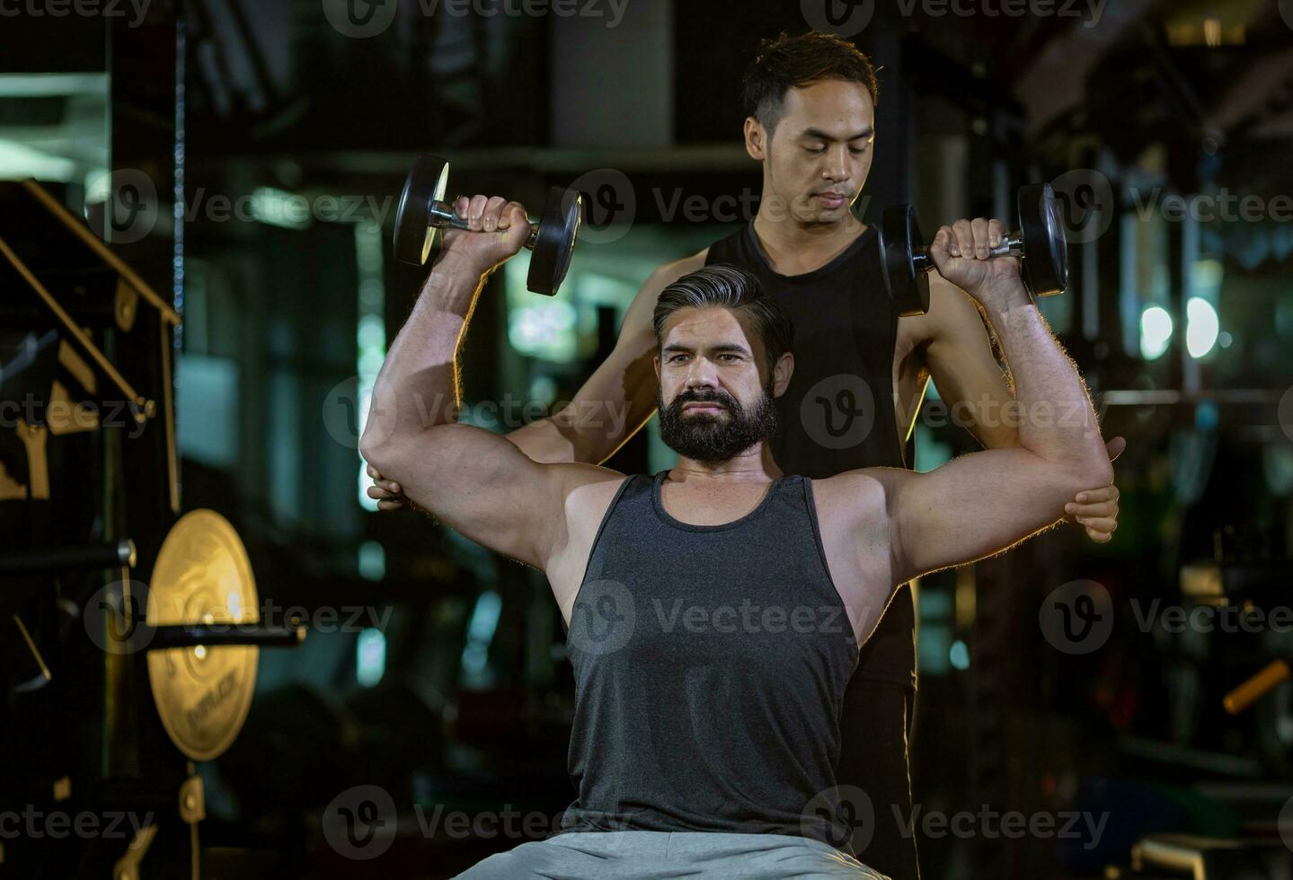 Caucasian man is using dumbbell in weight training for arm muscle inside gym with support from trainer to prevent injury dark background for exercising and workout concept photo