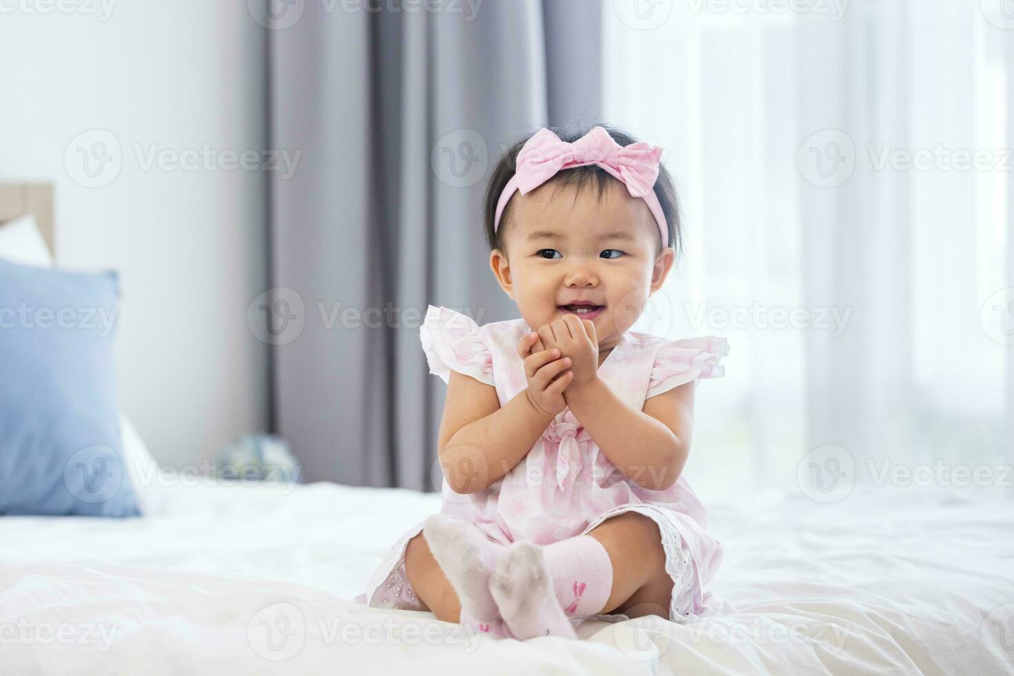 Asian baby toddler in cute pink dress is smiling while sitting on the bed with happiness for healthy kid and adorable girl portrait usage photo