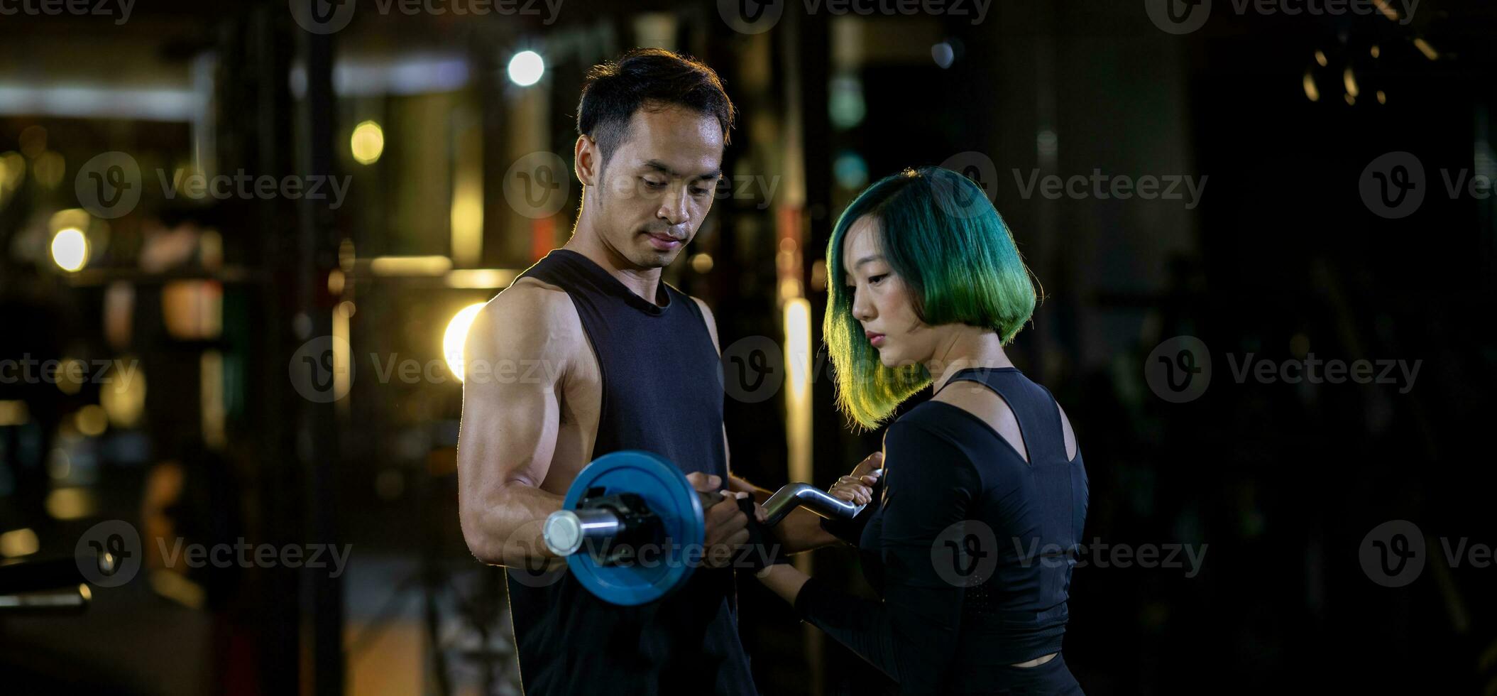 Asian woman is using easy bar as beginner in weight training on barbell for arm and core muscle inside gym with support from trainer to prevent injury dark background for exercising and workout photo