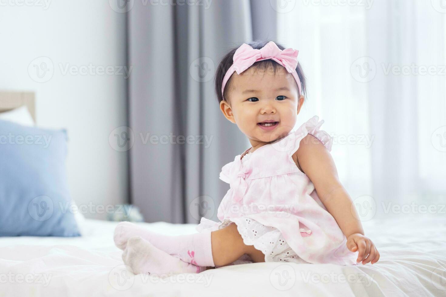 Asian baby toddler in cute pink dress is smiling while sitting on bed with happiness for healthy kid and adorable girl portrait concept photo